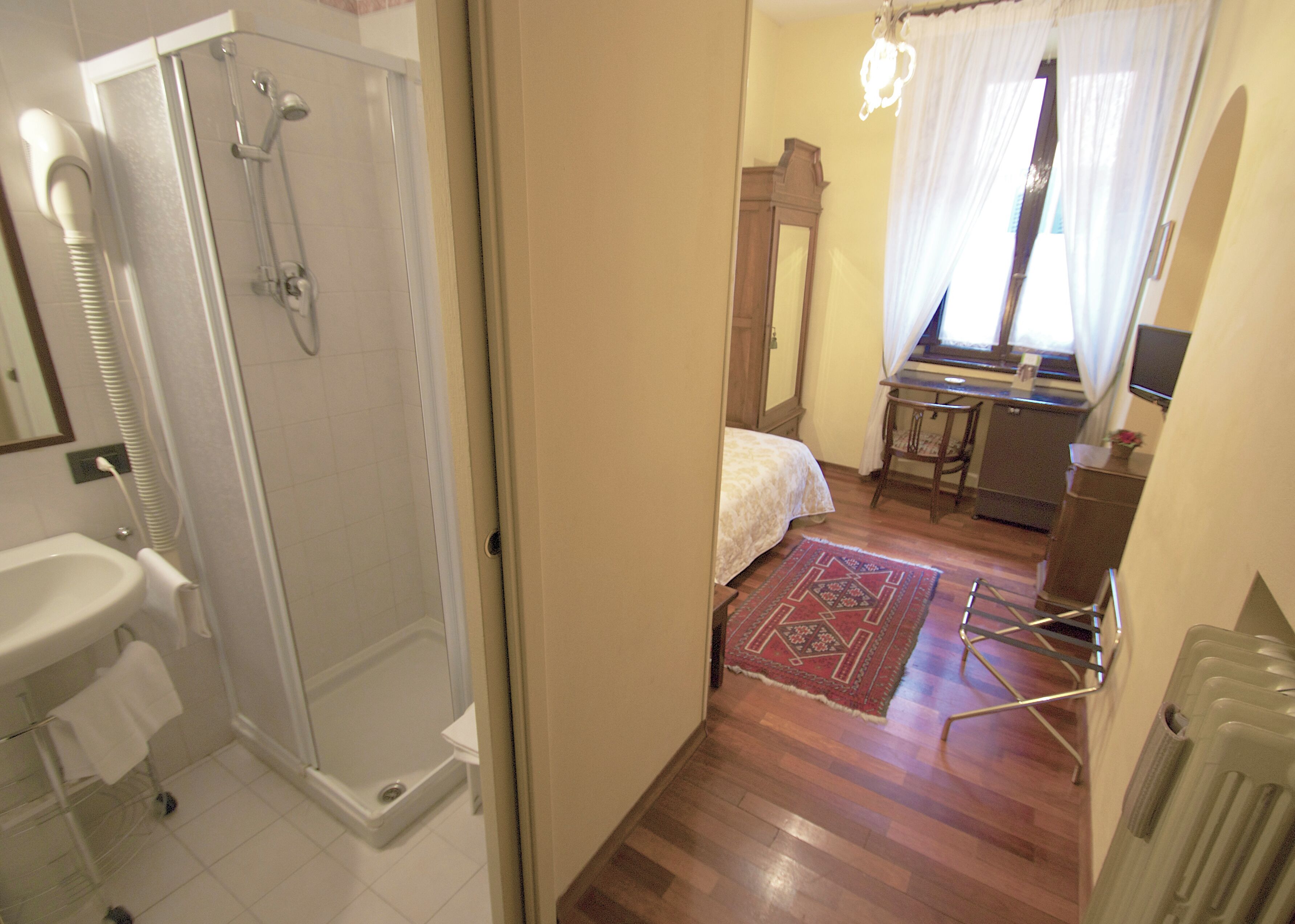 A bathroom with a shower, sink, and toilet. There is a bed with a nightstand and a dresser in the adjacent room.