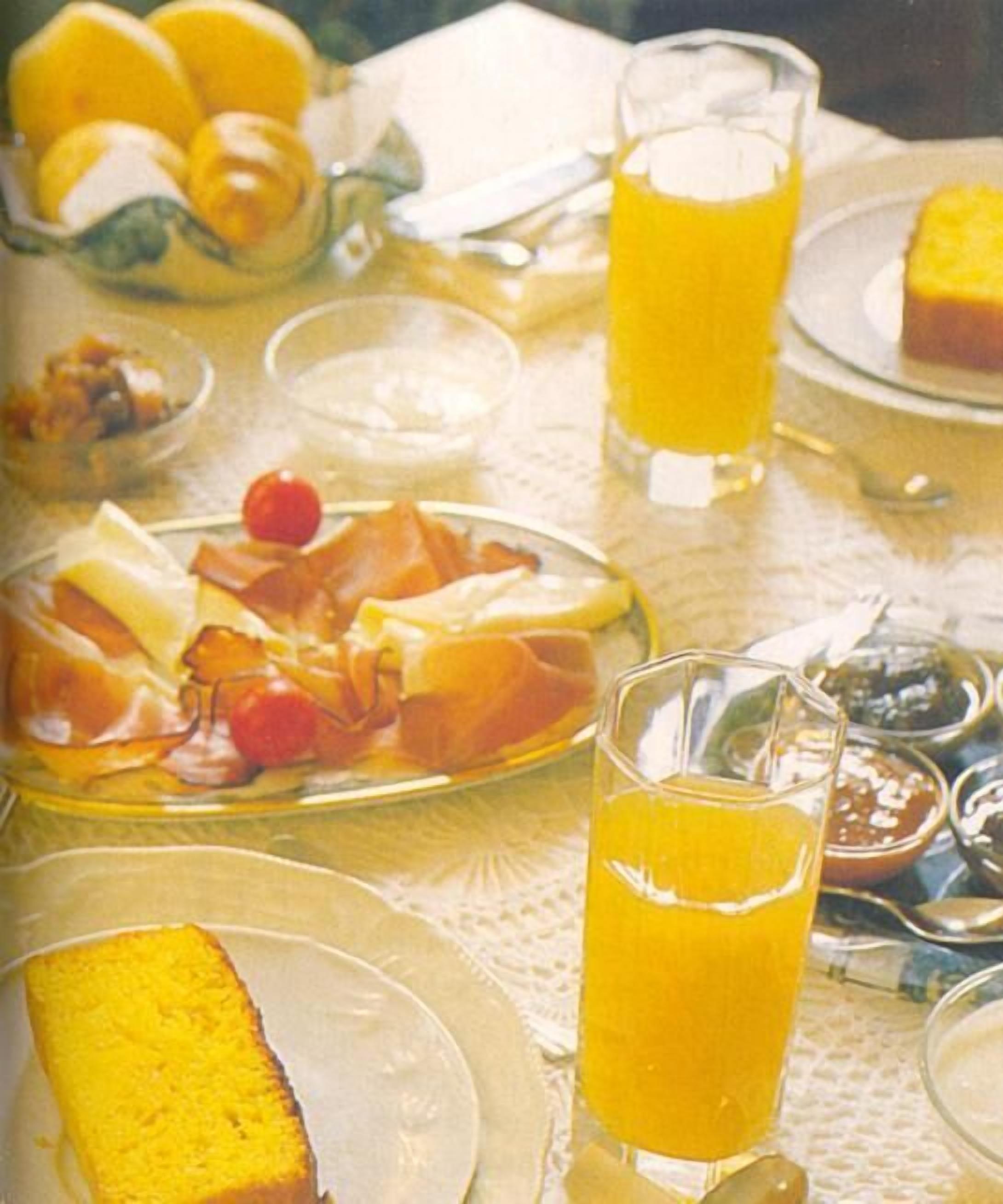 Une table mise pour le petit-déjeuner, avec des assiettes de nourriture, un verre de jus d’orange et un bol de fruits.