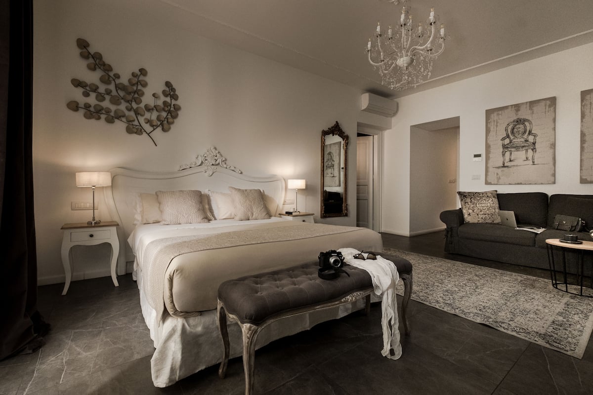 Un dormitorio con una cama grande, mesitas de noche, un banco, una araña y obras de arte en las paredes.
