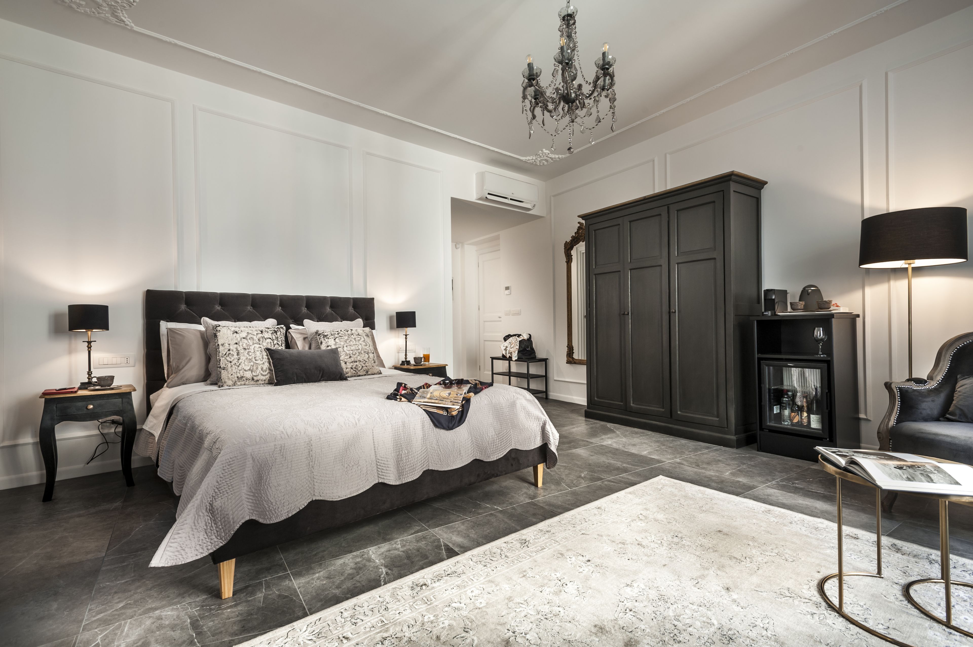 A bedroom with a large bed, a dark wardrobe, a chandelier, and a side table with a lamp.