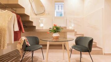 A modern room with a glass table, two chairs, a vase with flowers, a coat rack with clothes, and a window with a view of the outside.
