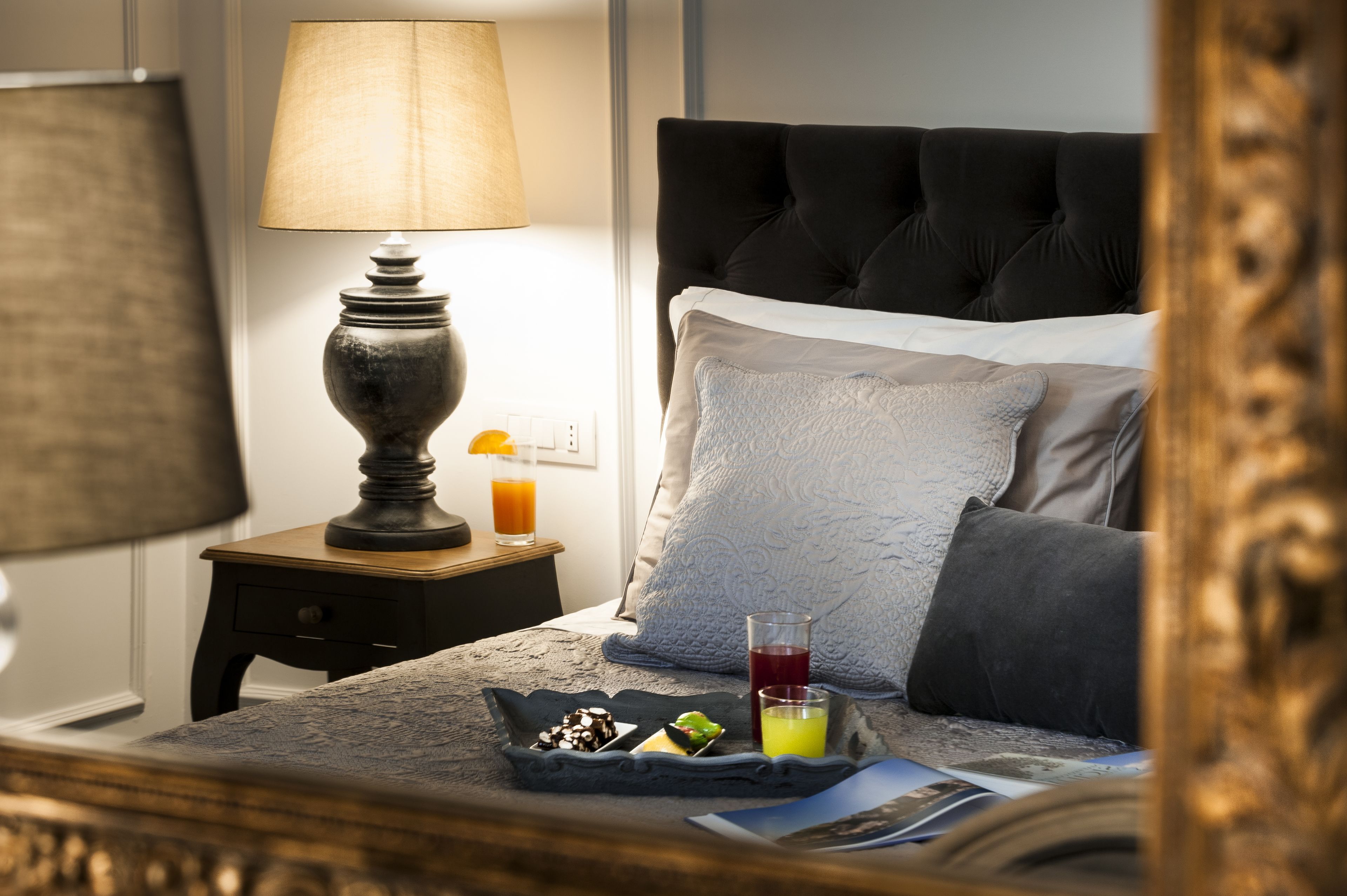 A neatly made bed with a grey headboard, a bedside table with a lamp, a tray with breakfast items, and a glass of orange juice.