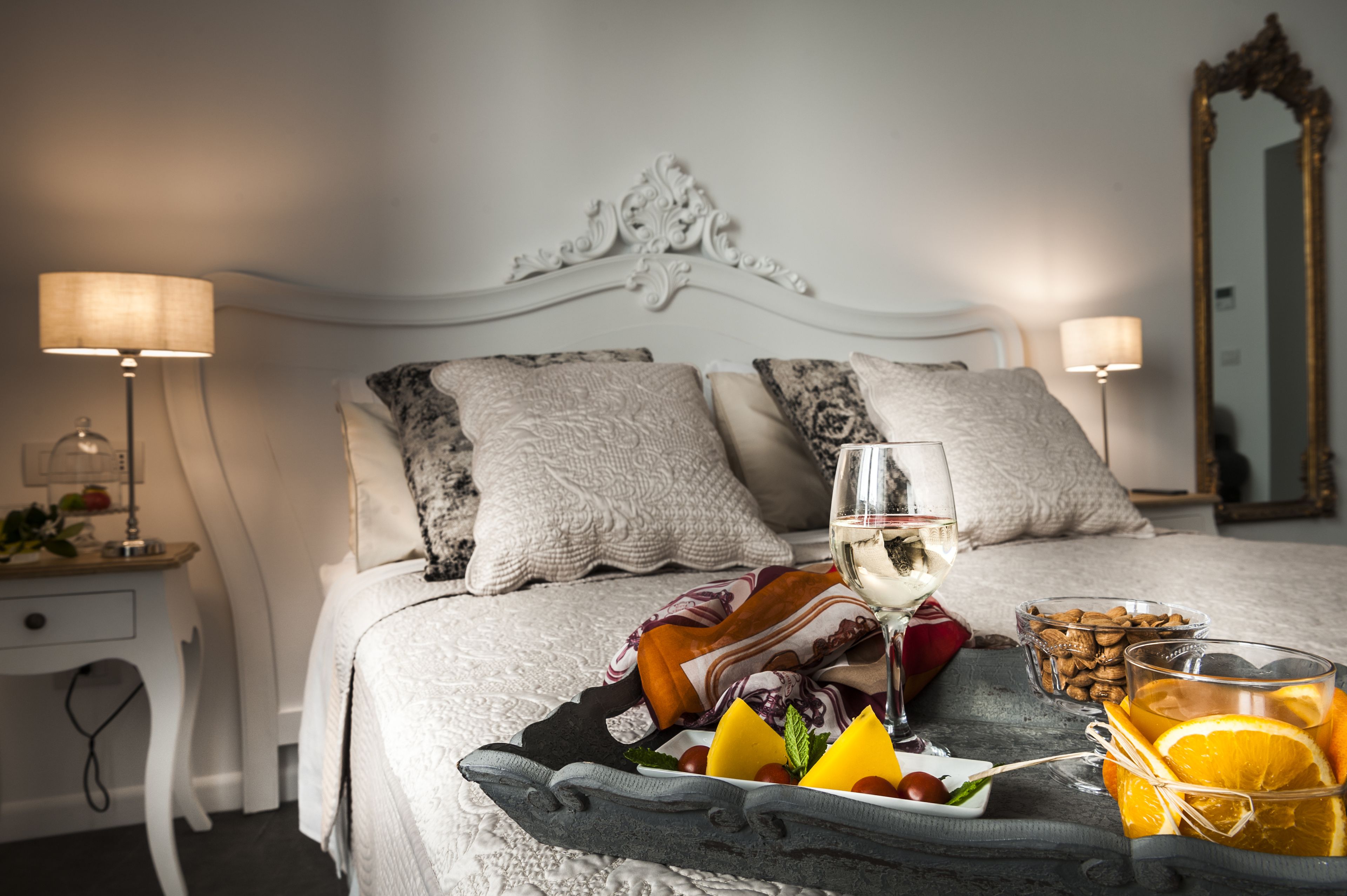 A neatly made bed with a tray of fruit and a glass of white wine, a bedside table with a lamp, and a mirror on the wall.