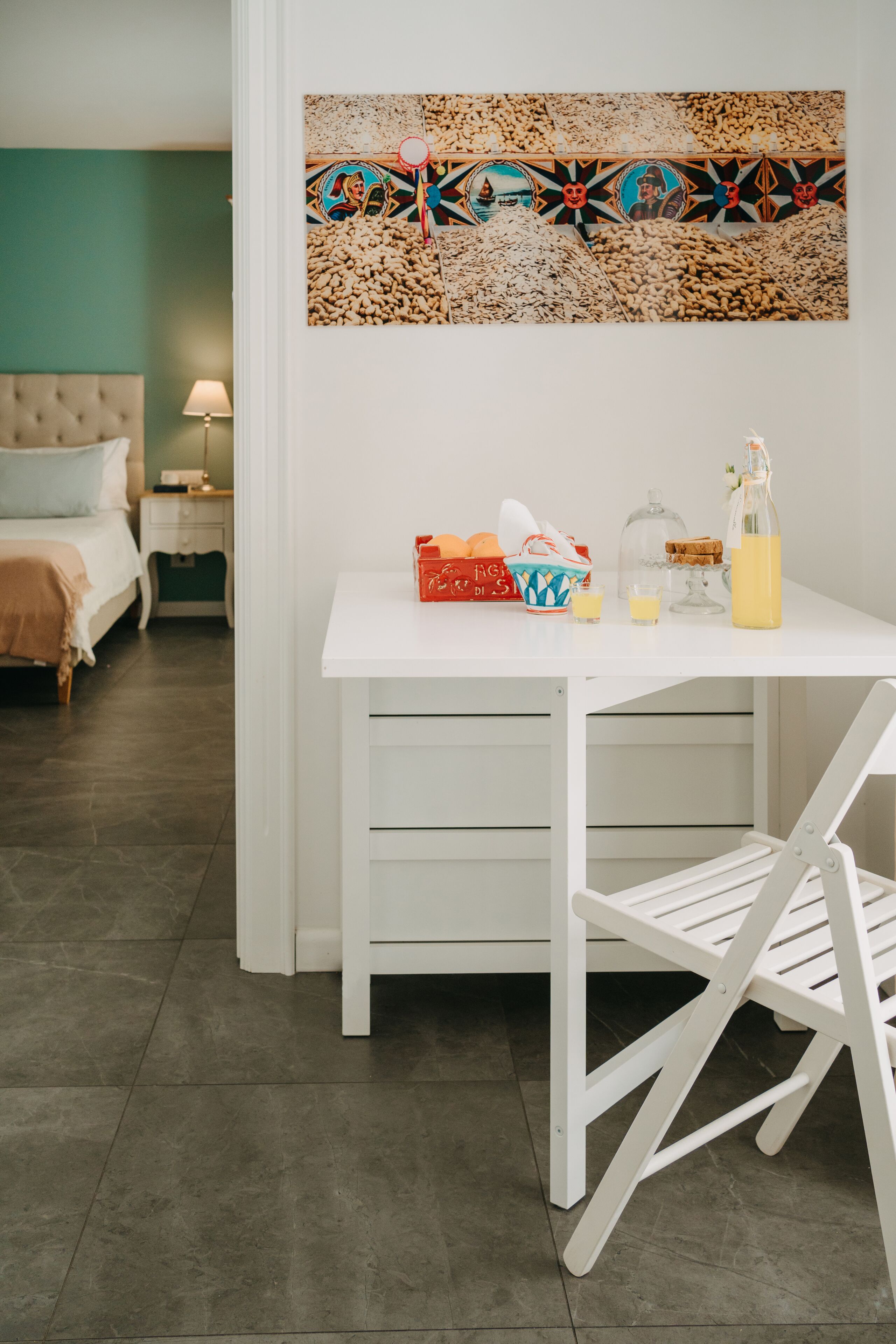 A neatly arranged room with a bed, bedside table, lamp, and a desk with a chair. A colorful abstract painting is on the wall above the desk.