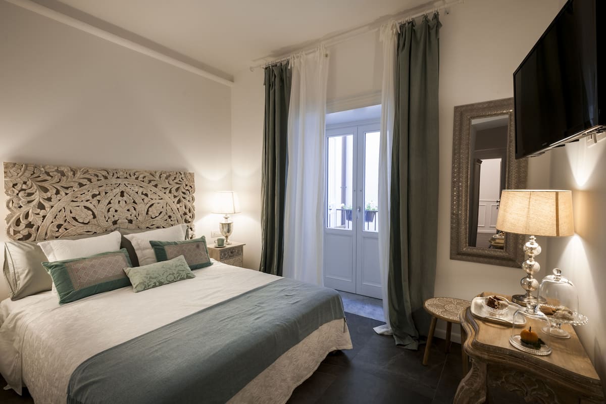 Un dormitorio ordenado con una cama grande, una mesita de noche con lámpara, una pequeña mesa con lámpara y adornos, y un espejo en la pared.