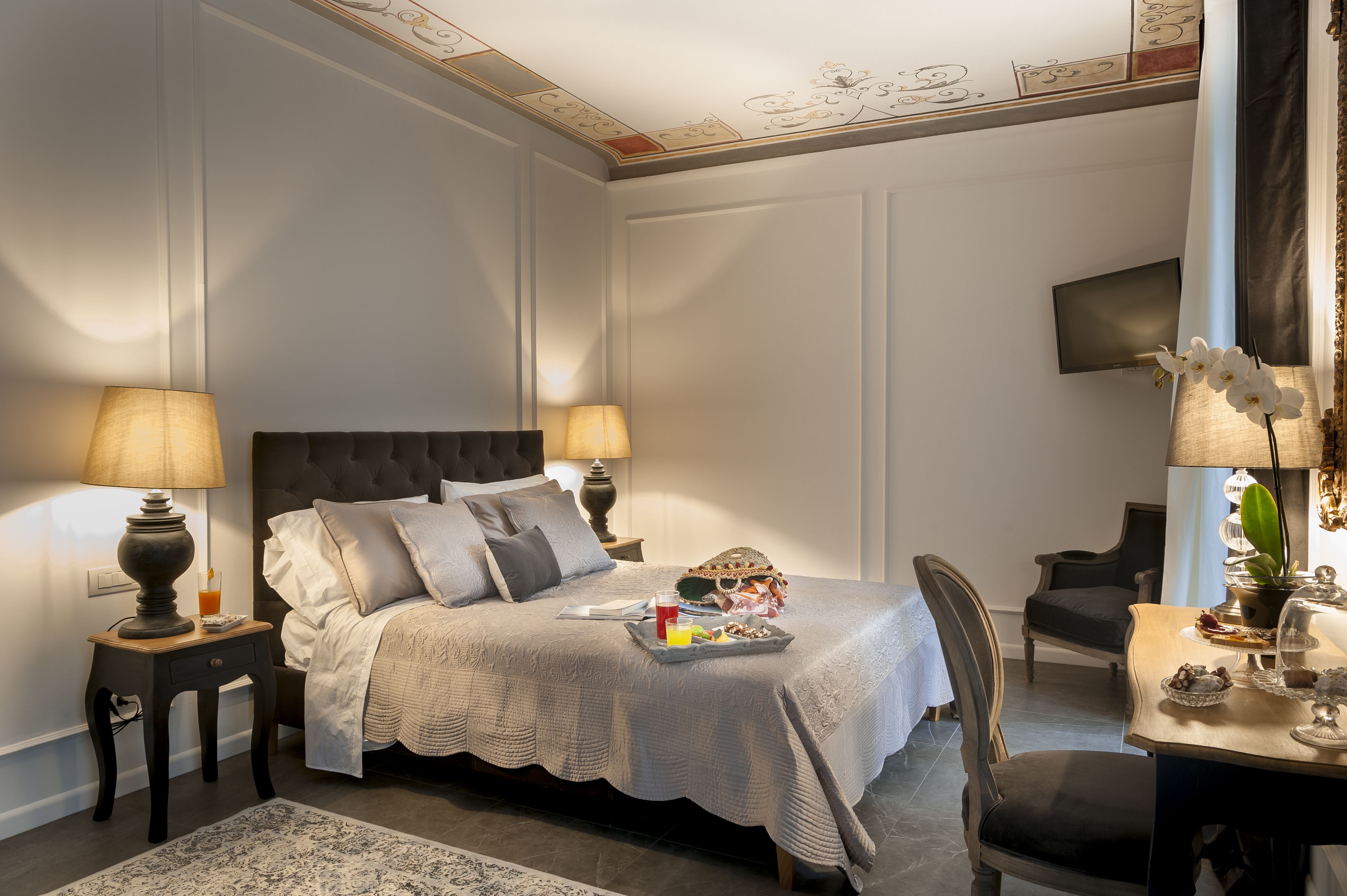 A neatly arranged bedroom with a large bed, bedside lamps, a tray with breakfast items, and a desk with a chair.