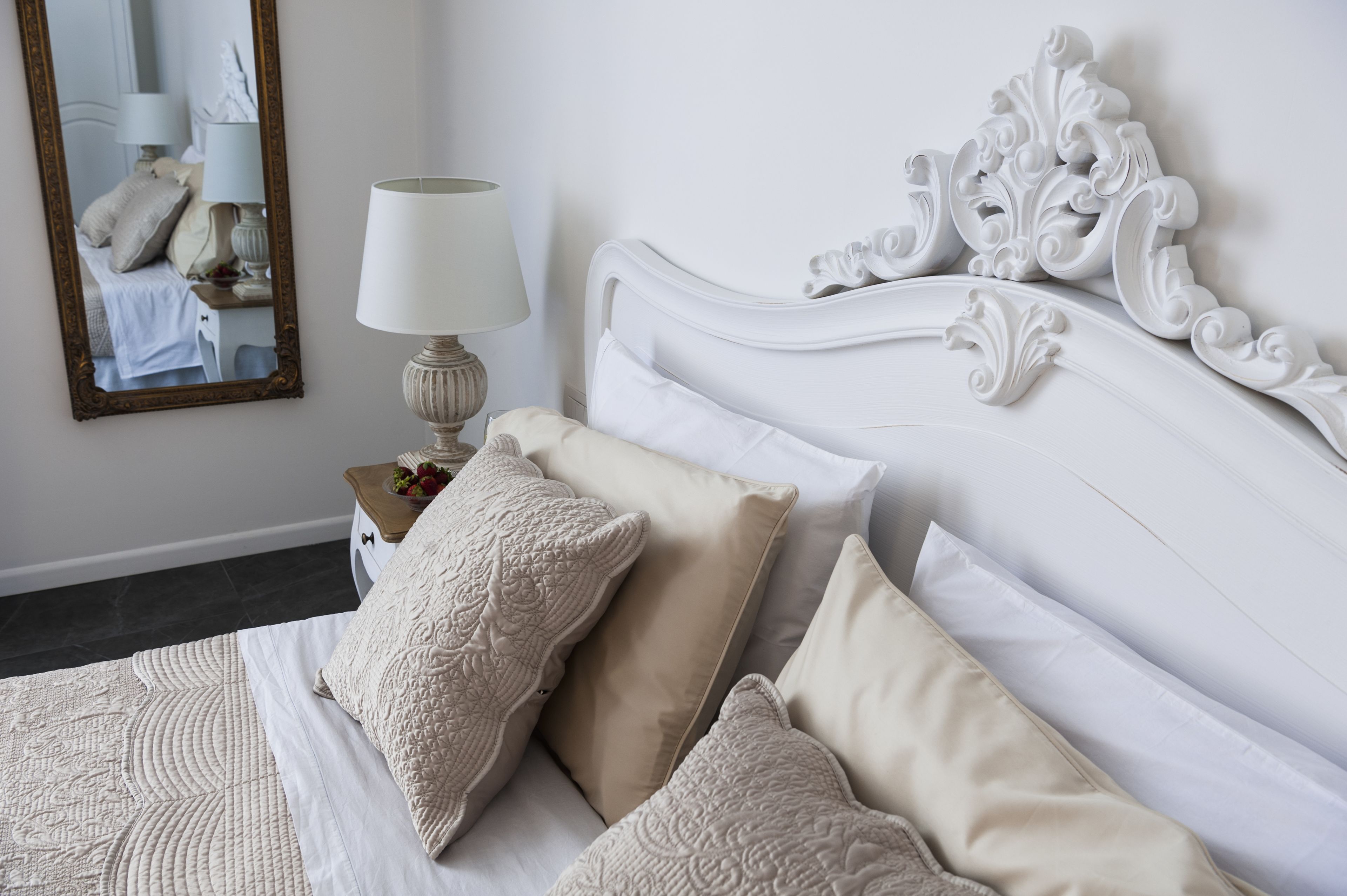 A neatly made bed with a white headboard and several pillows, a bedside lamp, and a mirror on the wall.