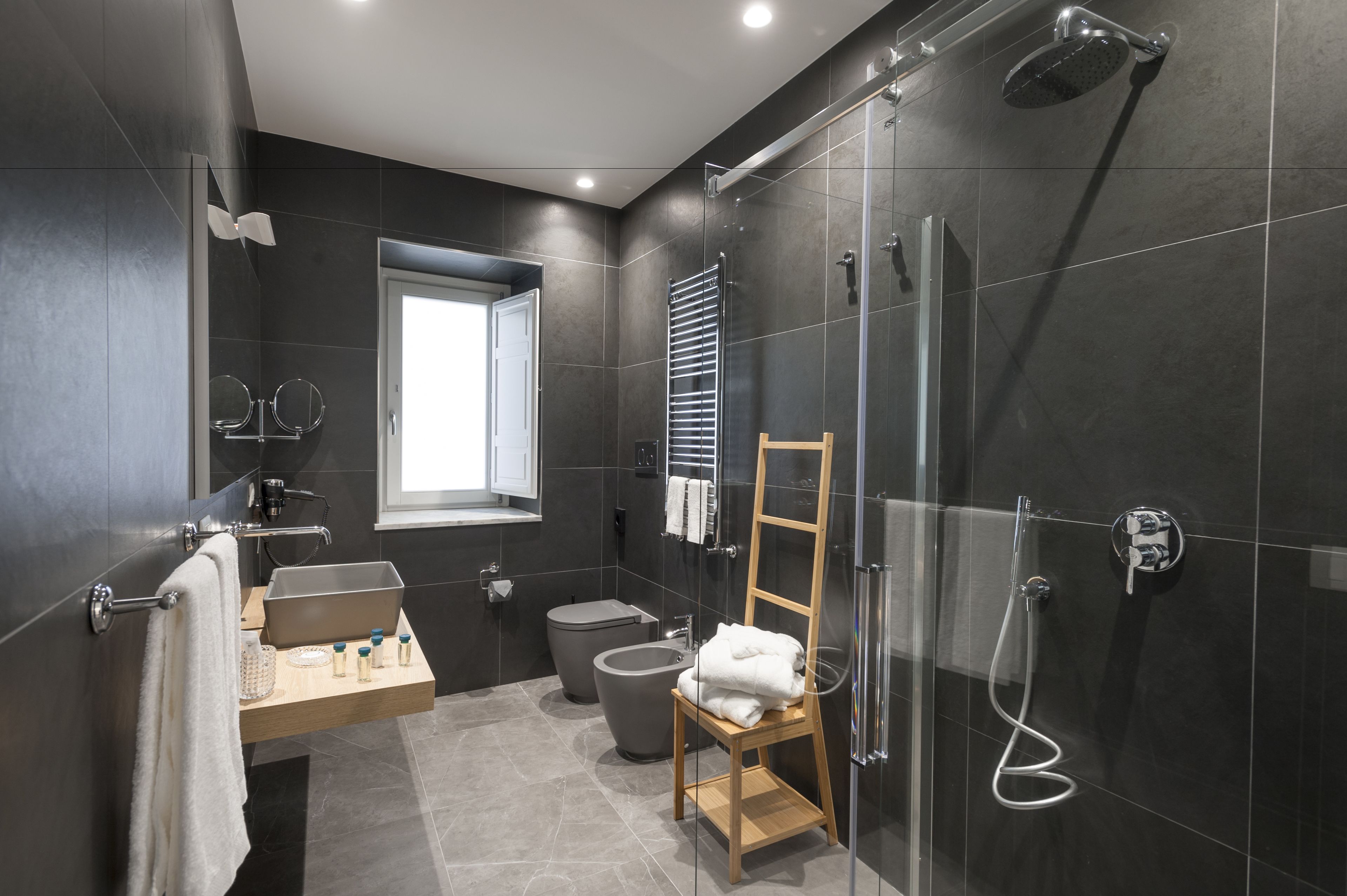 Un baño moderno con cabina de ducha de vidrio, una silla de madera, un lavabo con espejo y una ventana que proporciona luz natural.