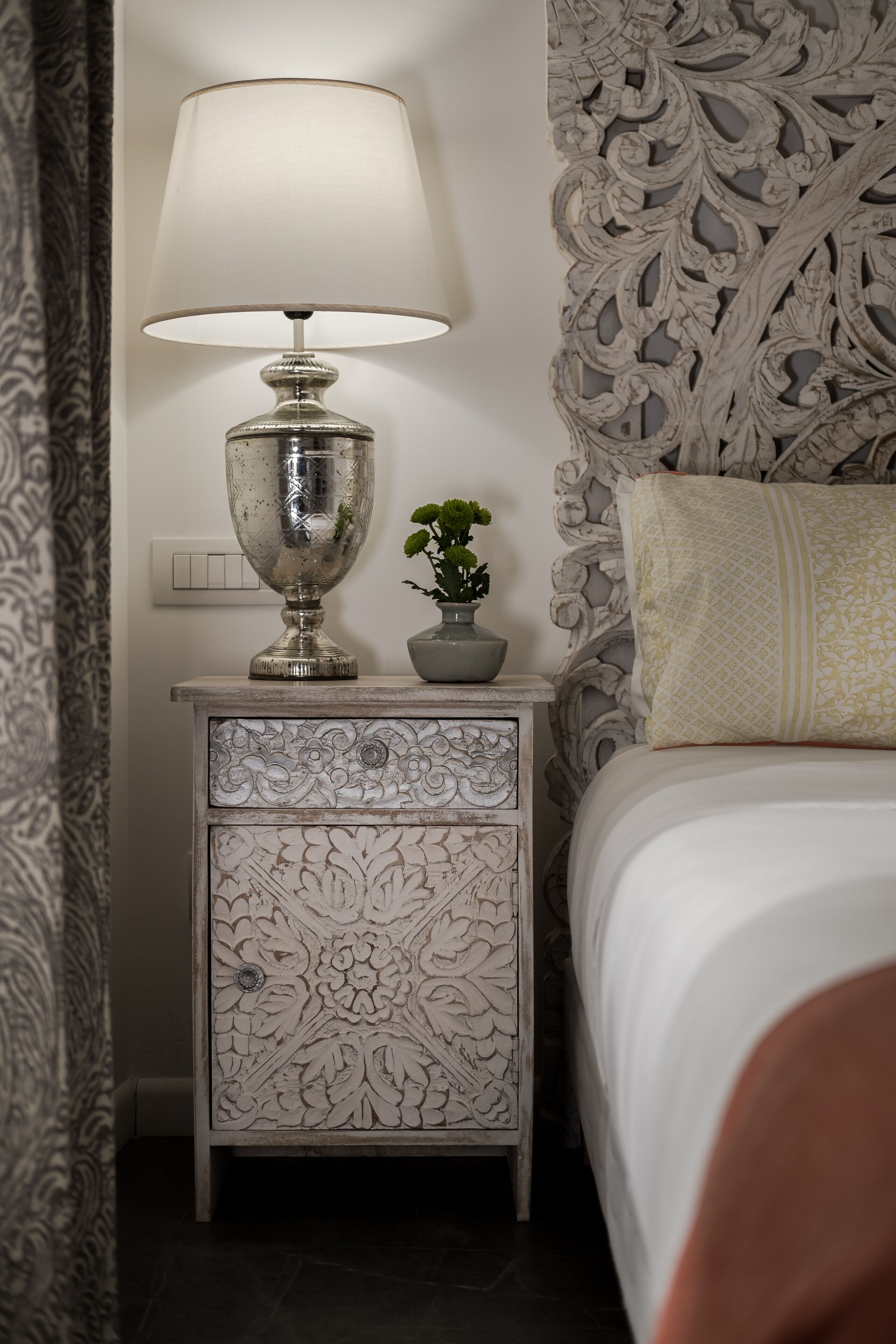 A neatly made bed with a white comforter and a decorative pillow. A silver lamp with a white shade is placed on a carved wooden nightstand beside the bed. A small potted plant is also on the nightstand.