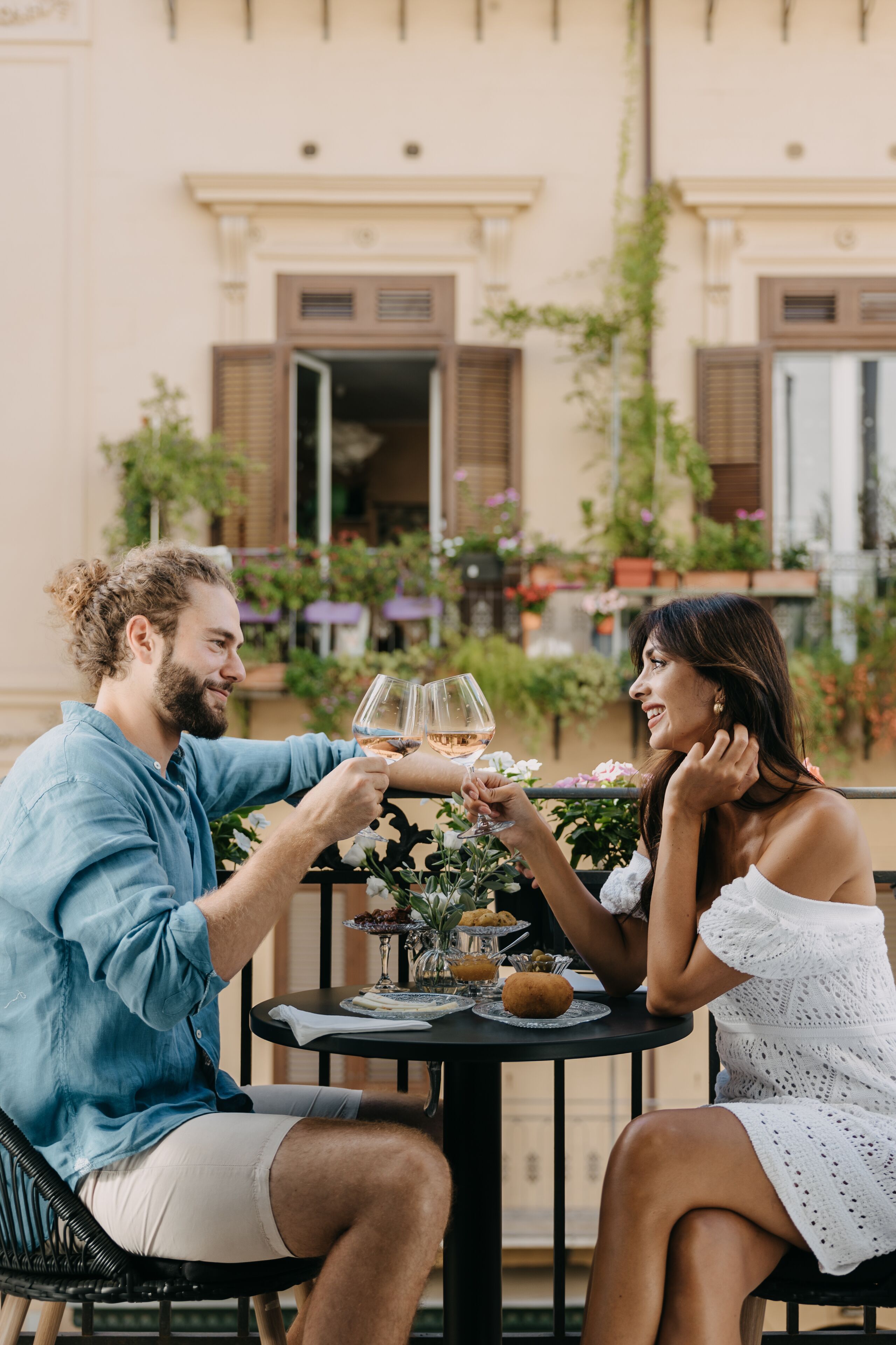 A couple toasting with wine glasses at an outdoor table with a floral centerpiece, surrounded by greenery and a building facade.