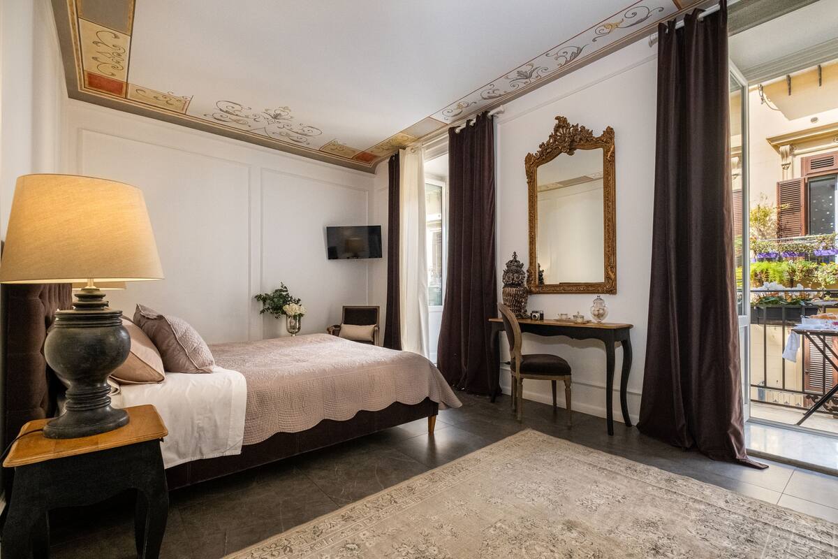 Un dormitorio con cama, escritorio, silla, lámpara, espejo y vista al balcón exterior.