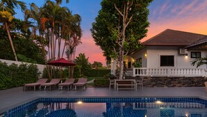 A swimming pool with lounge chairs and a red umbrella, surrounded by palm trees and a house with a tiled roof. - Happy Family Villa 2  (Chalong)