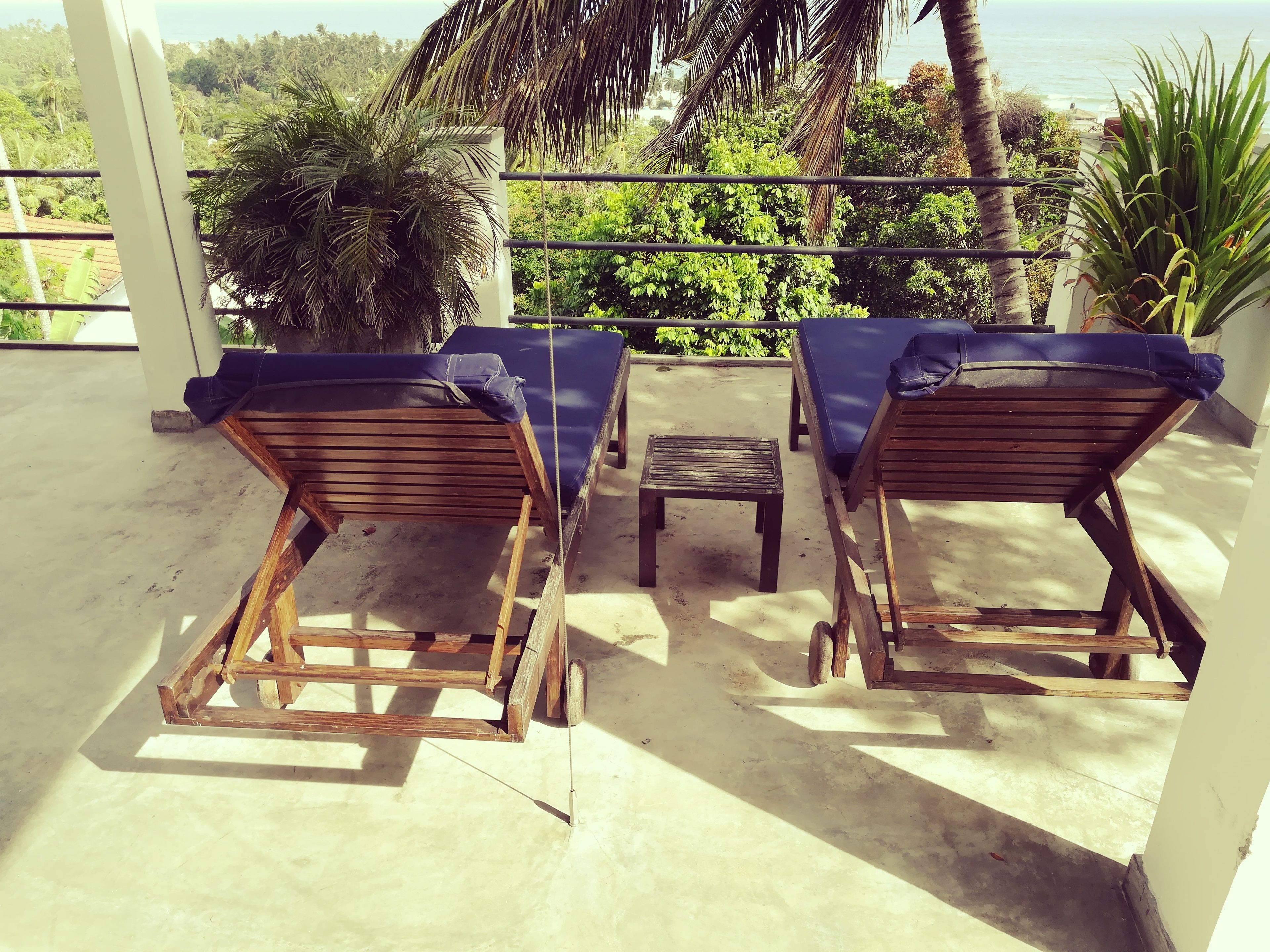 Two wooden lounge chairs with blue cushions on a balcony, a small table, and a view of the ocean and greenery.