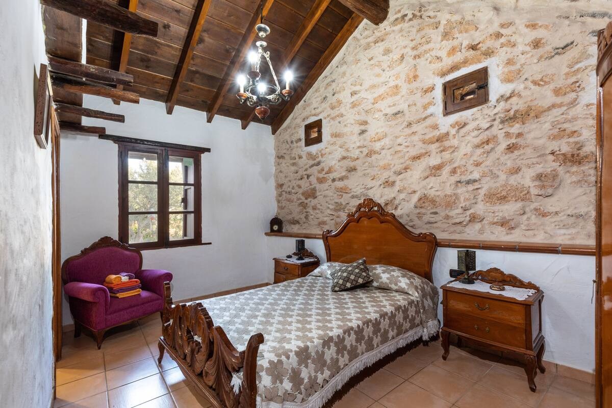 Ein Schlafzimmer mit einem hölzernen Bett, einem Nachttisch, einem Sessel und einer Steinwand.