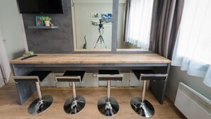 Ein modernes Zimmer mit großem Schreibtisch, drei Barhocker, einem an der Wand montierten Fernseher und einem Stativ.