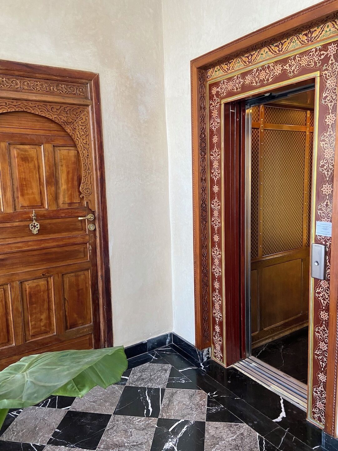 Une porte en bois ornée de sculptures complexes et dotée d’une poignée en métal, un sol en marbre à carreaux et un mur à motifs.