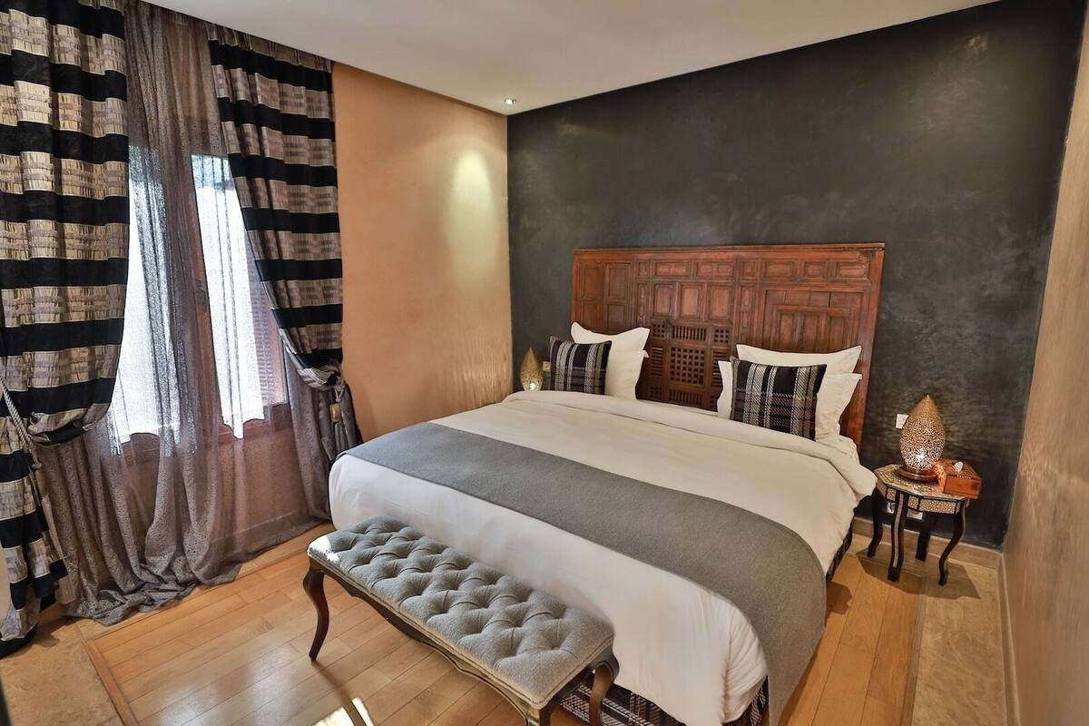 Quarto com uma cama grande, cabeceira de madeira, um banquinho e uma mesa de cabeceira com abajur.