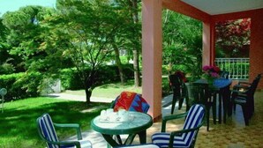 A covered outdoor seating area with chairs and a table, surrounded by lush greenery and a tiled floor. - Lignano Riviera Spacious (Lignano Sabbiadoro)