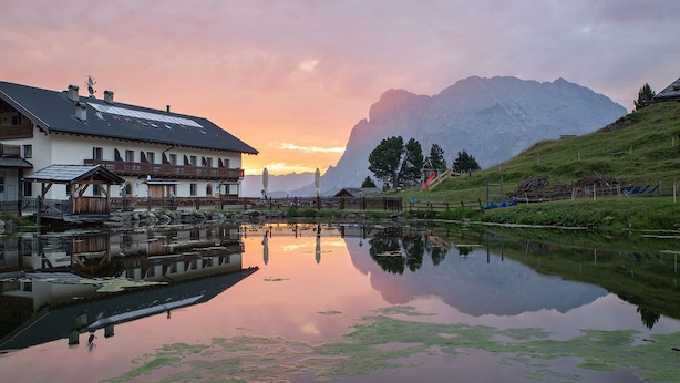 A traditional chalet with a reflection in a pond, surrounded by mountains and a clear sky.