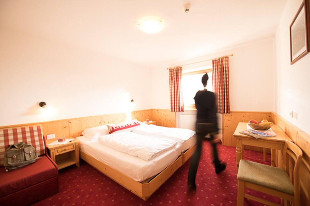 Une chambre d’hôtel avec un lit en bois, une petite table de chevet, une chaise, un canapé rouge et une fenêtre avec des rideaux.