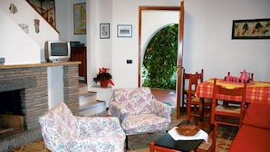 A living room with a fireplace, patterned armchairs, a dining table with a red tablecloth, and a view of greenery through an archway. - Villa in Lignano Riviera Comfortable (Lignano Sabbiadoro)