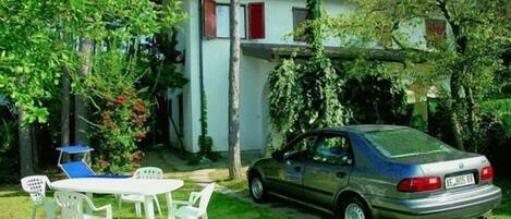 A car parked in a grassy area with a white building in the background, surrounded by trees and greenery.