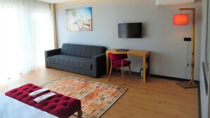 A modern hotel room with a grey sofa, a small wooden desk, a red chair, a TV mounted on the wall, and a large window with sheer curtains. - VVR Hotel (Istanbul)