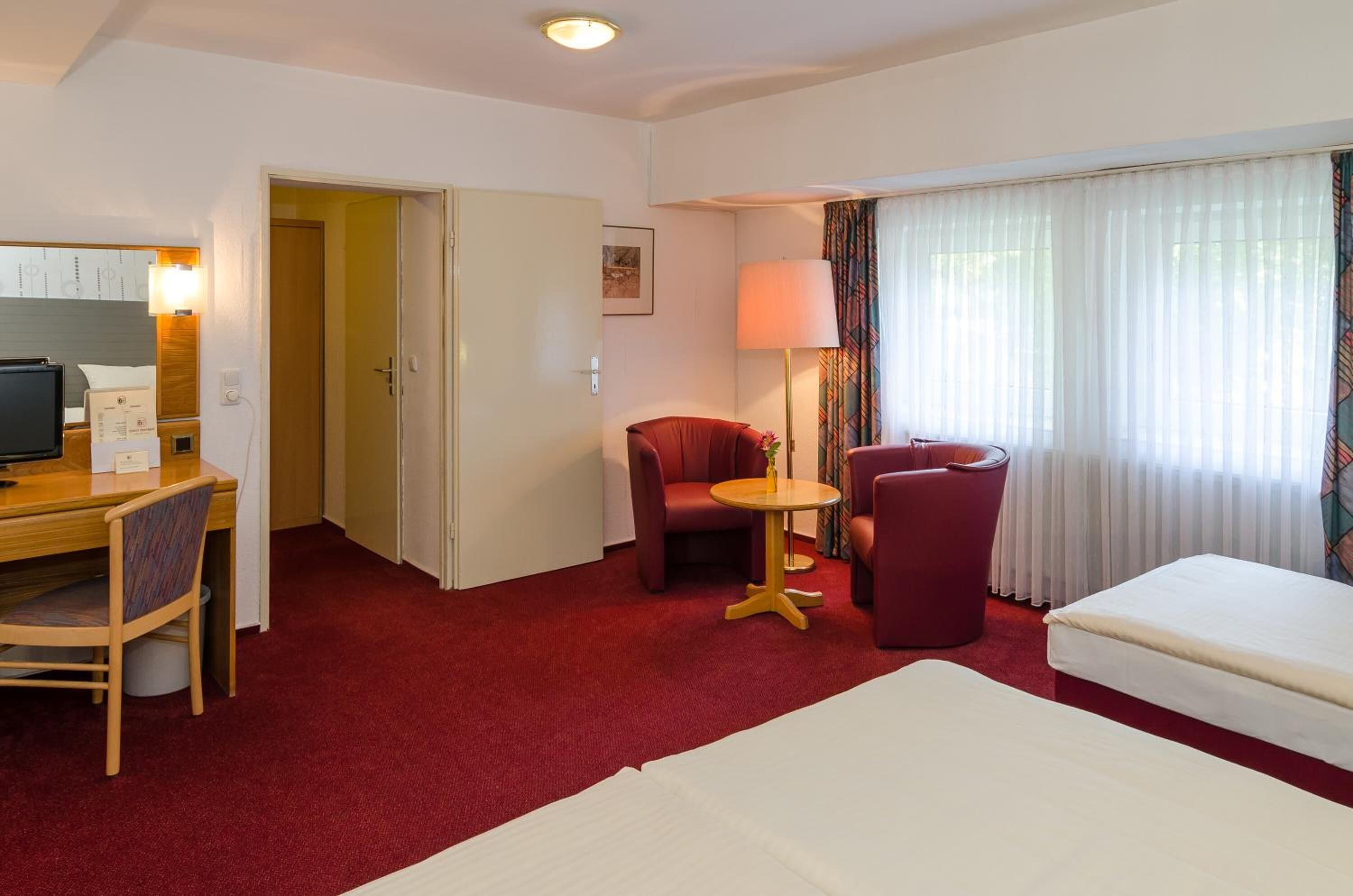 Een hotelkamer met een bed, een bureau met stoel, een klein rond tafeltje met lamp, en een rode fauteuil.