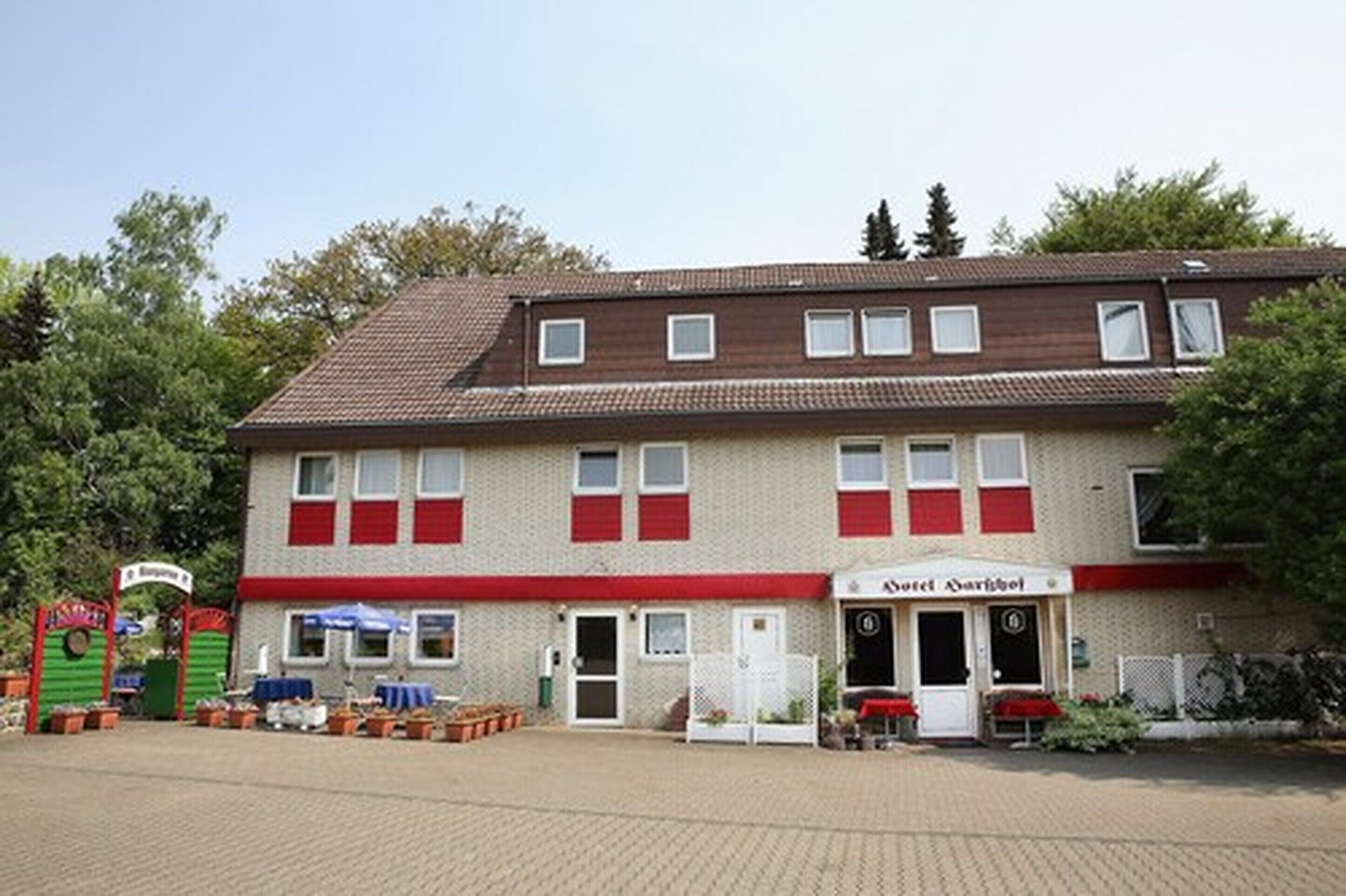 A two-story building with a red and white facade, a sign for 'Hotel Amisberg' on the ground floor, and a paved entrance area with potted plants.