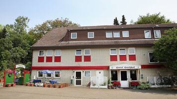 A two-story building with a red and white facade, a sign for 'Hotel Amisberg' on the ground floor, and a paved entrance area with potted plants.