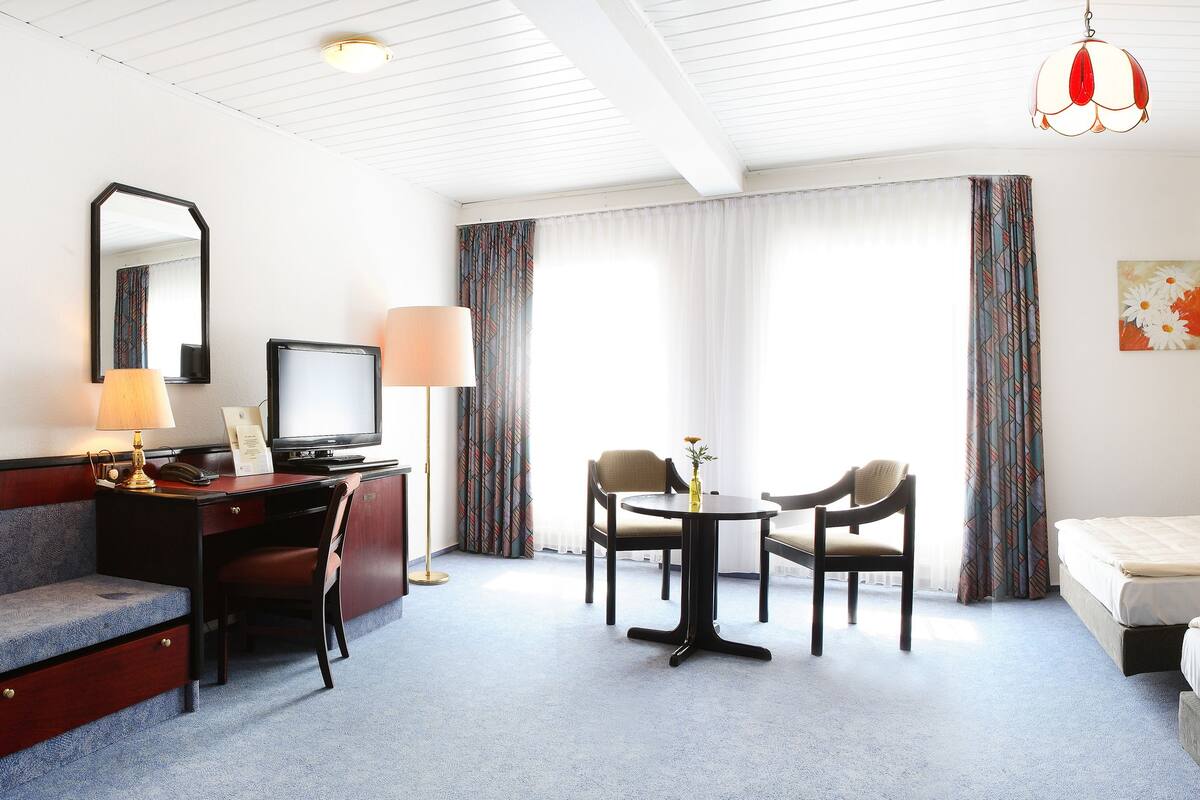 A hotel room with a desk, two chairs, a television, a bed, and a bedside table.