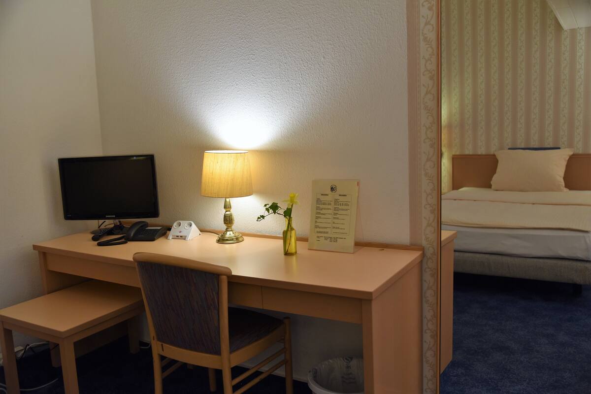A hotel room with a desk, chair, computer, lamp, and a bed.