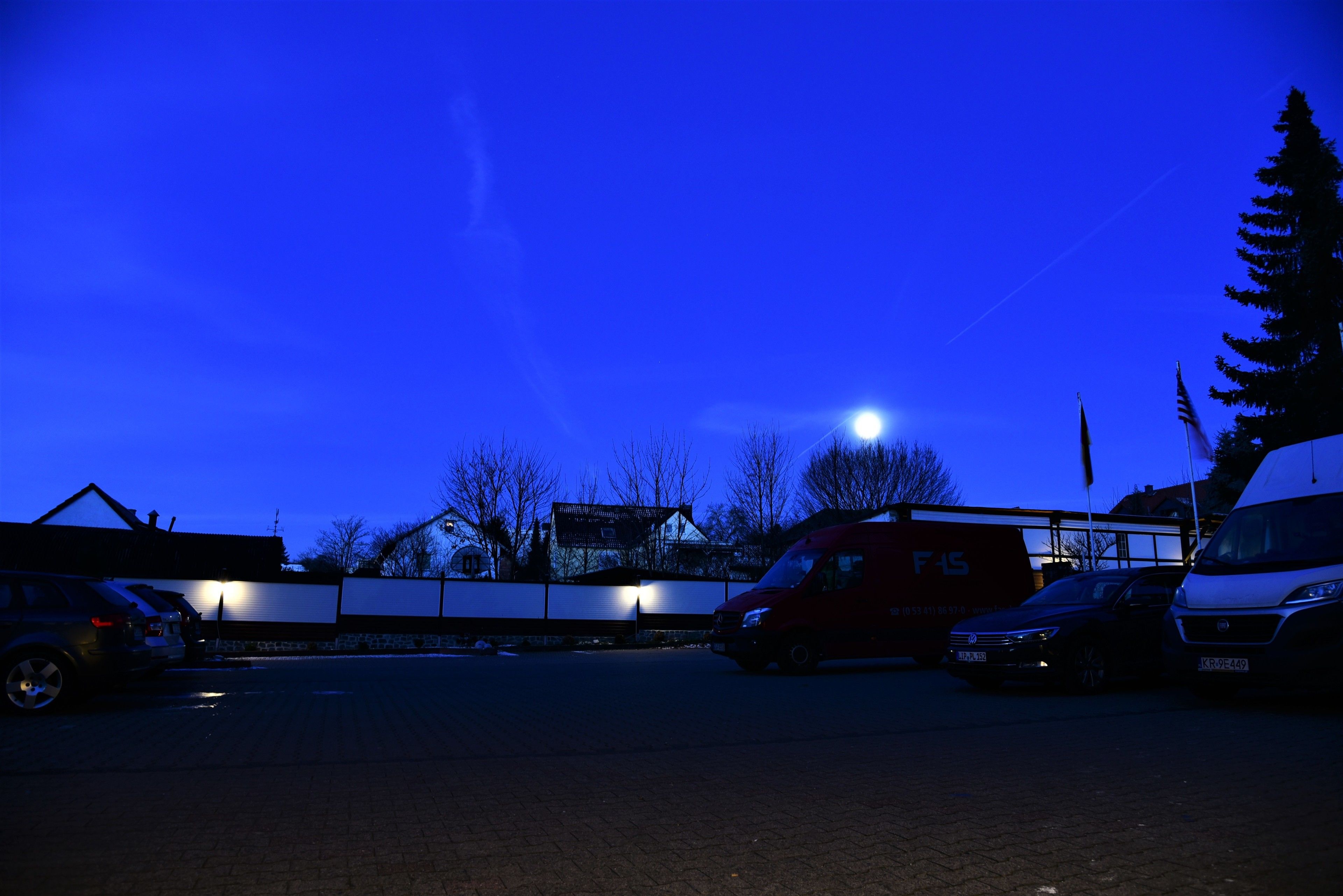 Aparcamiento al anochecer con varios vehículos, incluyendo una camioneta roja y una camioneta blanca, estacionados bajo un cielo despejado con una luna visible.