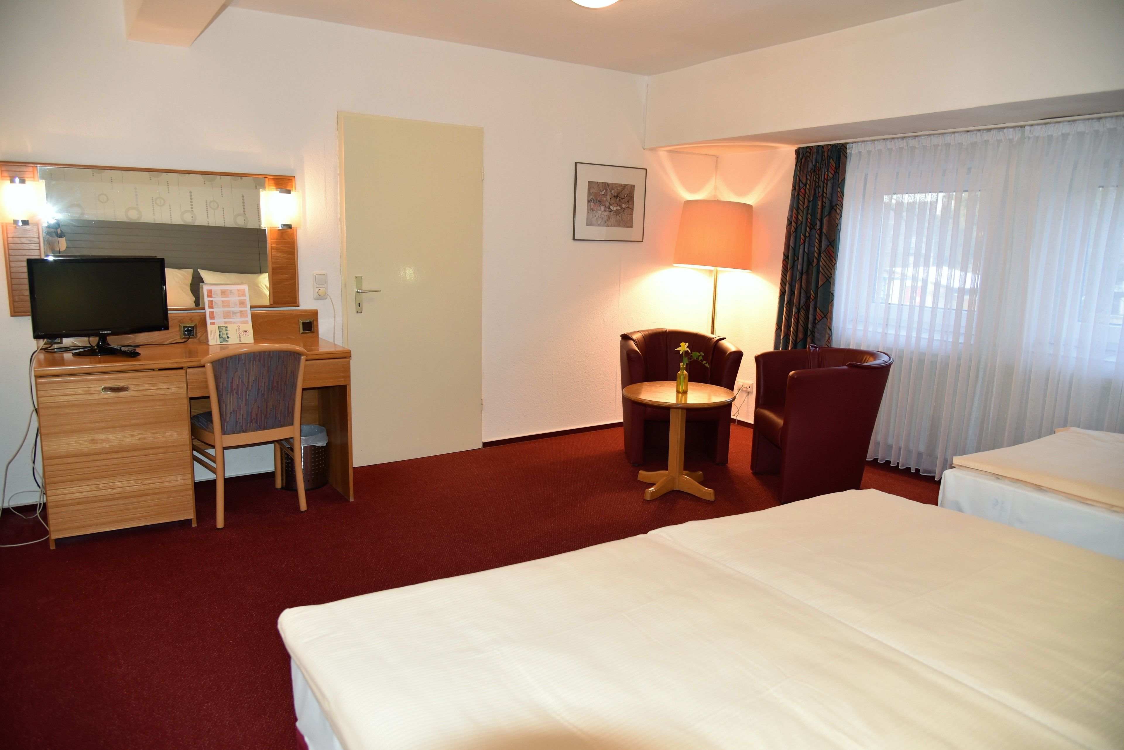Habitación de hotel con una cama, un escritorio con computadora, dos sillas, una lámpara y una ventana con cortinas.