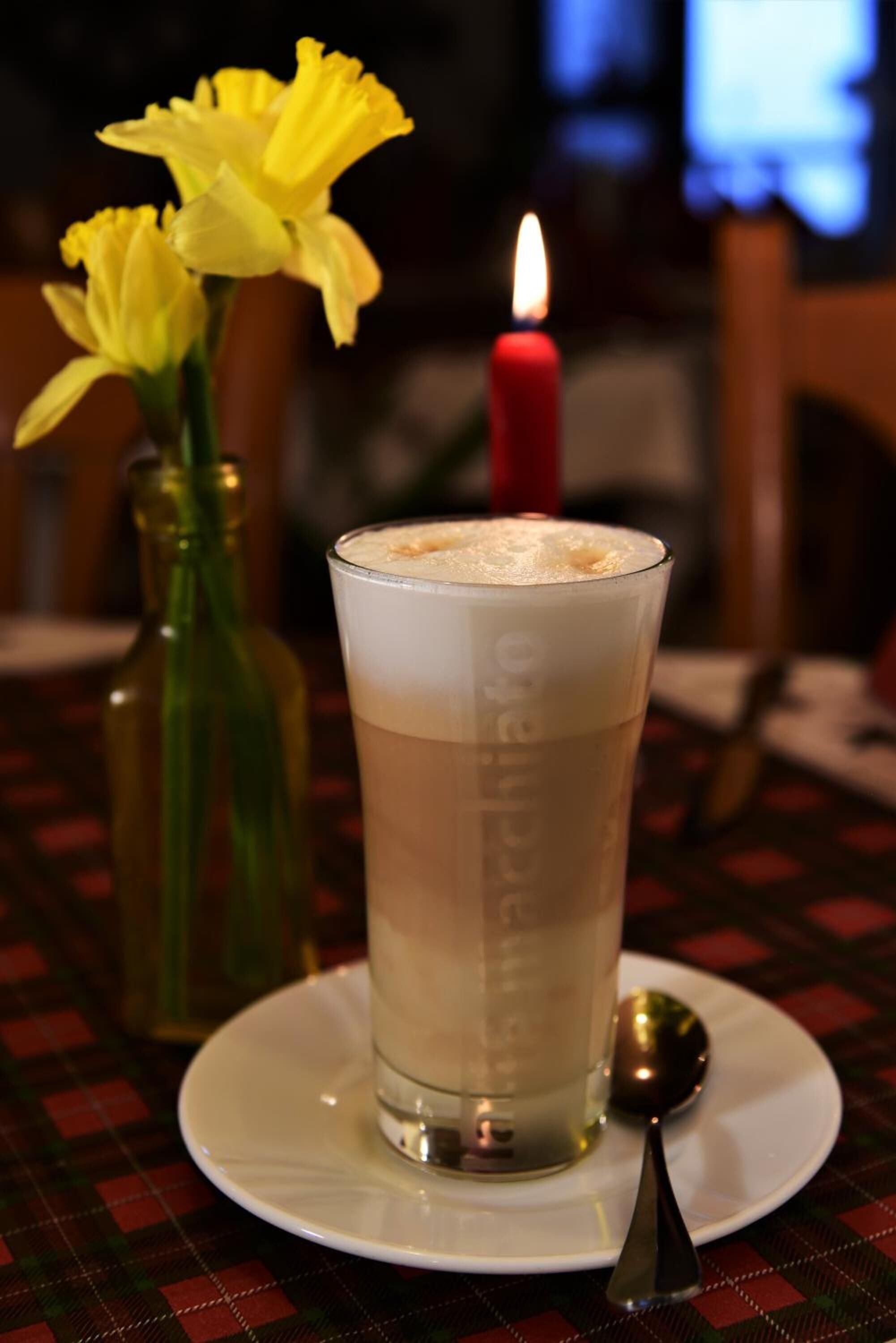Un vaso de una bebida de color claro con espuma, sobre un plato blanco, junto a una cuchara. Se ve una vela encendida al fondo, y una botella sobre la mesa.