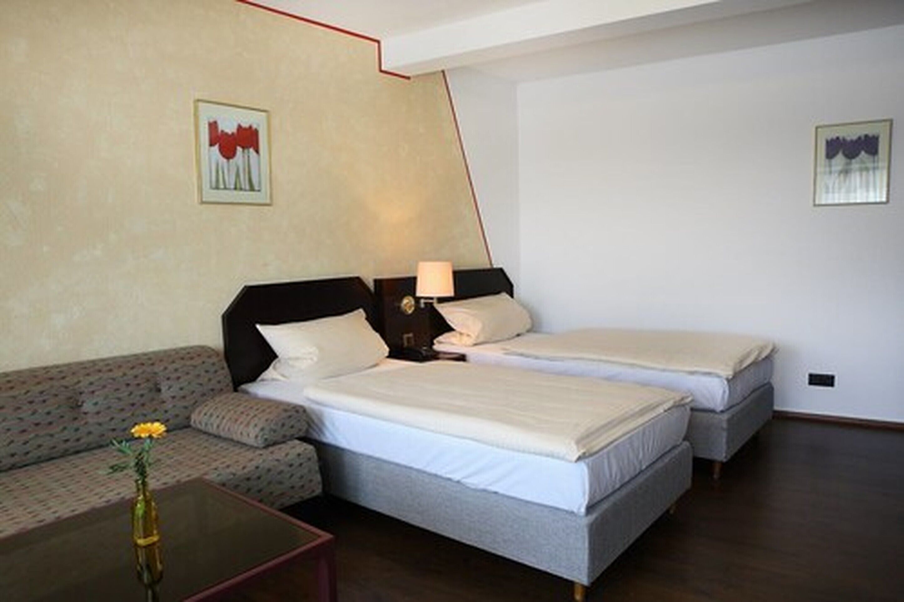 Ein Hotelzimmer mit einer Couch, zwei Einzelbetten, einem Nachttisch mit Lampe sowie einer Vase mit Blumen.