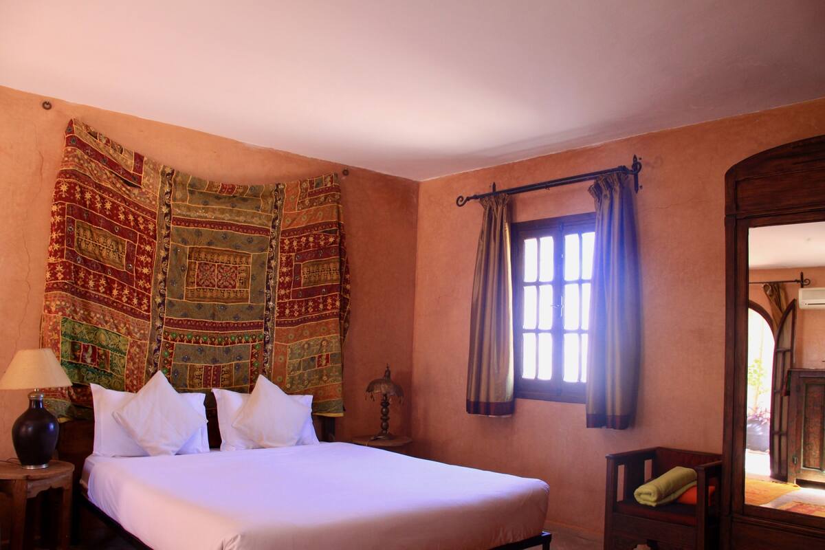Una habitación con una cama grande, una mesita de noche con lámpara, una silla, un ventanal con cortinas y un tapiz en la pared.
