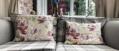 A sofa with floral cushions and a plaid pattern, positioned in a room with a brick wall and a window.
