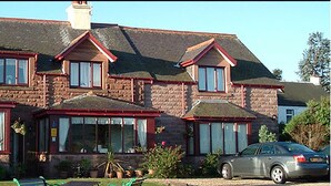 Une maison à deux étages avec une façade en pierre, de nombreuses fenêtres et une voiture garée dans l’allée.