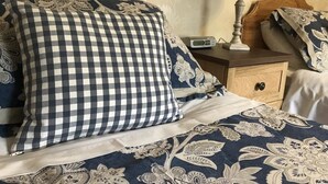 Un lit bien fait, avec un oreiller à carreaux, une couverture à motifs floraux et une table de chevet sur laquelle se trouvent une lampe et une radio.