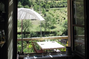 A balcony with a table and chairs, overlooking a green landscape with trees and a white umbrella.