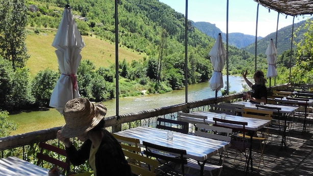 An outdoor dining area with tables and chairs, overlooking a river and green hills.