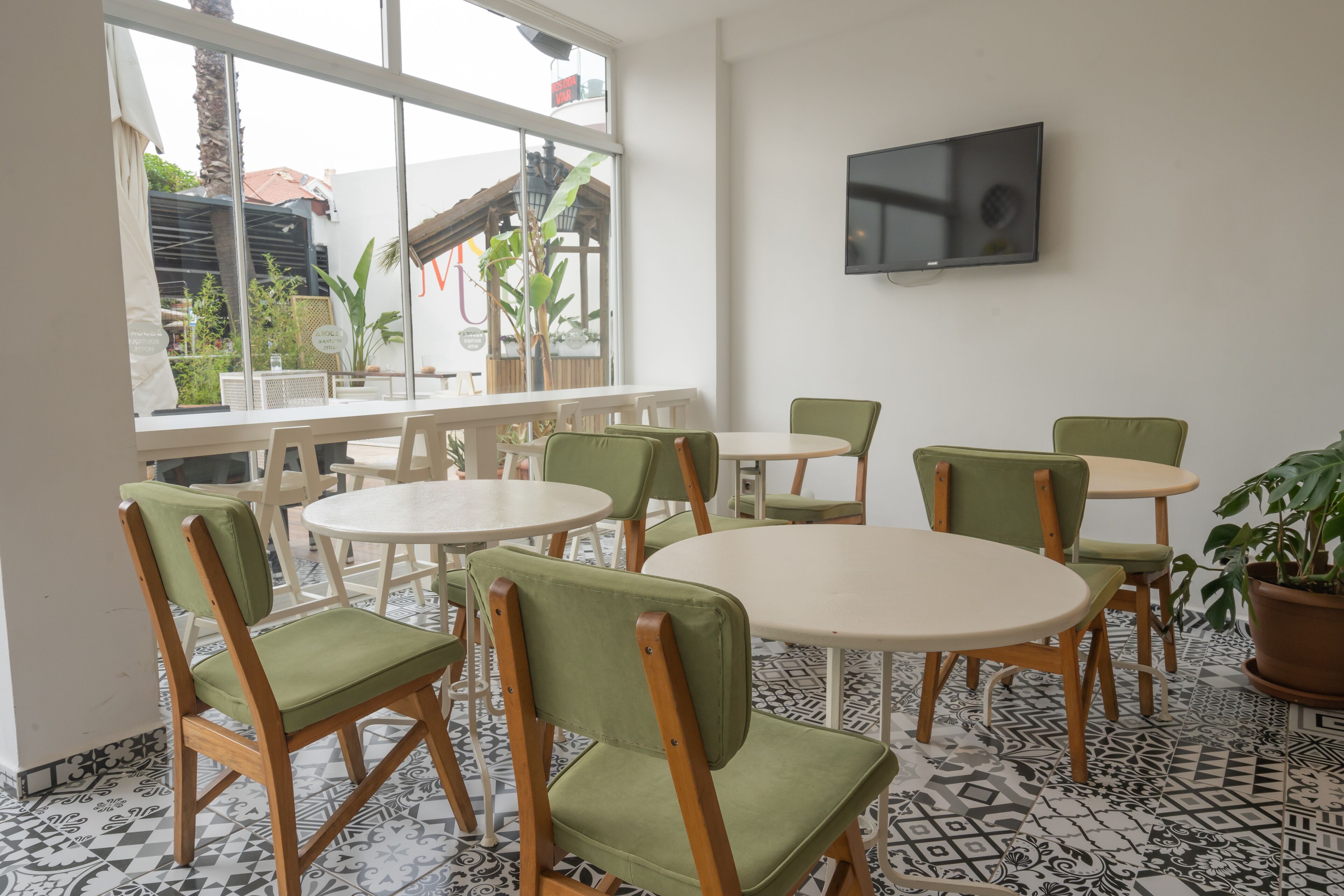 A dining area with round tables and green cushioned chairs, a TV mounted on the wall, and a view of the outdoors through large windows.