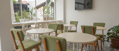 A dining area with round tables and green cushioned chairs, a TV mounted on the wall, and a view of the outdoors through large windows.