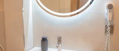 A bathroom with a white vanity, a round mirror with a light, a handheld hair dryer, and a towel rack.