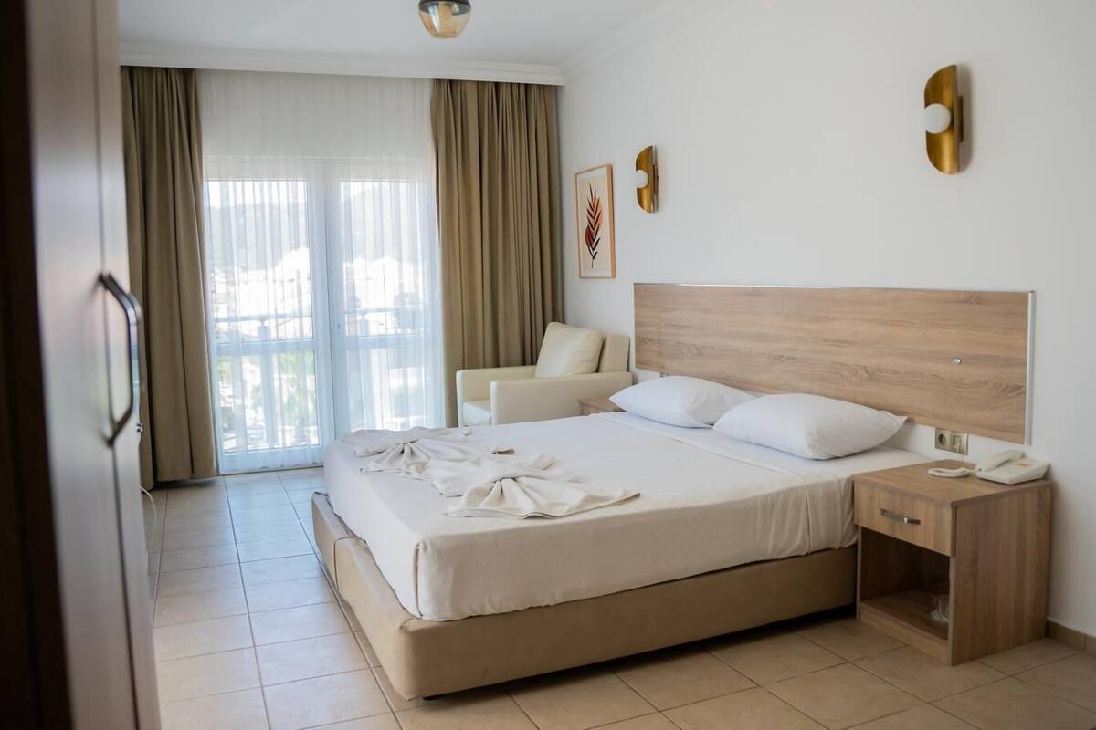 Ein Hotelzimmer mit einem großen Bett, einem Nachttisch, einem Sessel und Blick durch große Fenster nach draußen.