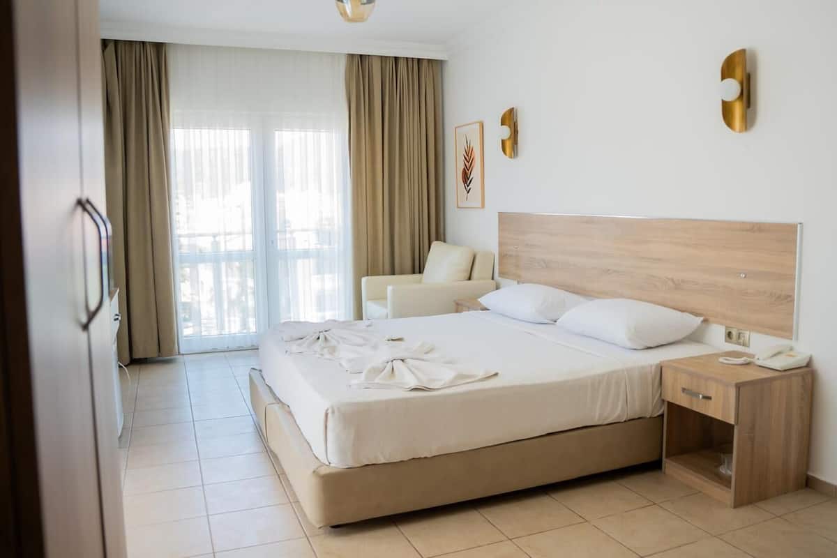 Ein Hotelzimmer mit einem großen Bett, einem Nachttisch, einem Sessel und Blick ins Freie durch das Fenster.