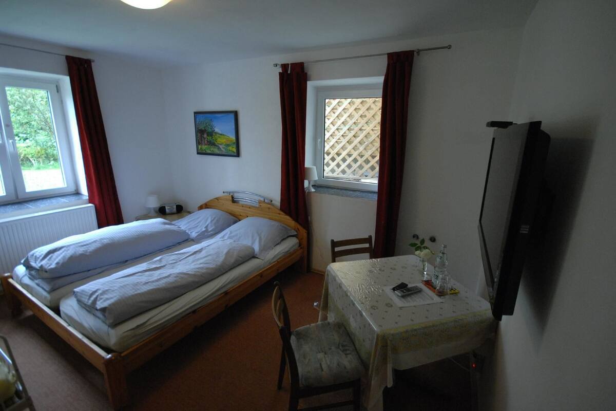 A bedroom with a double bed, a small table with a chair, a TV, and a window with curtains.