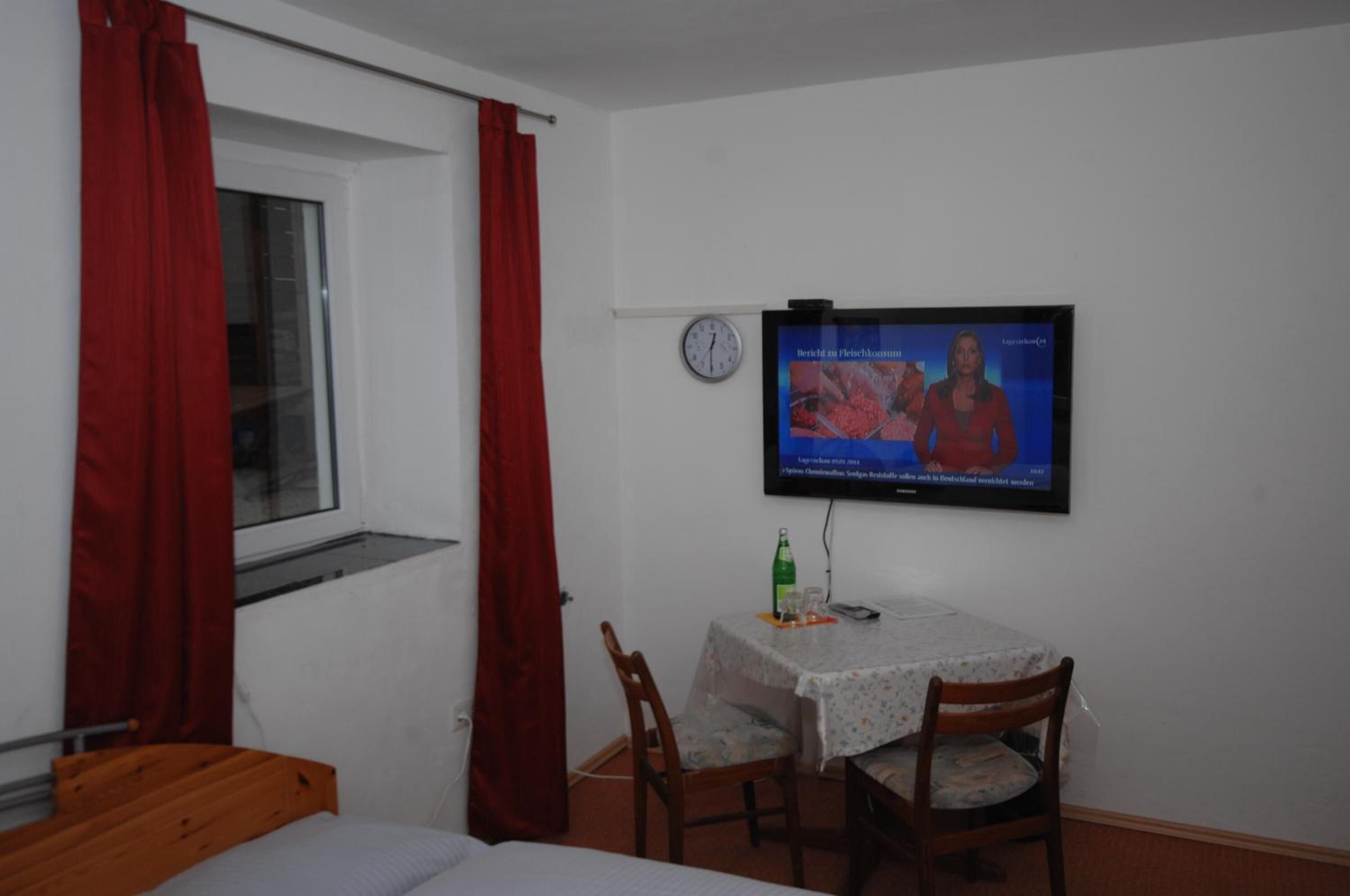 A small room with a bed, a table with a TV, and a window with red curtains.
