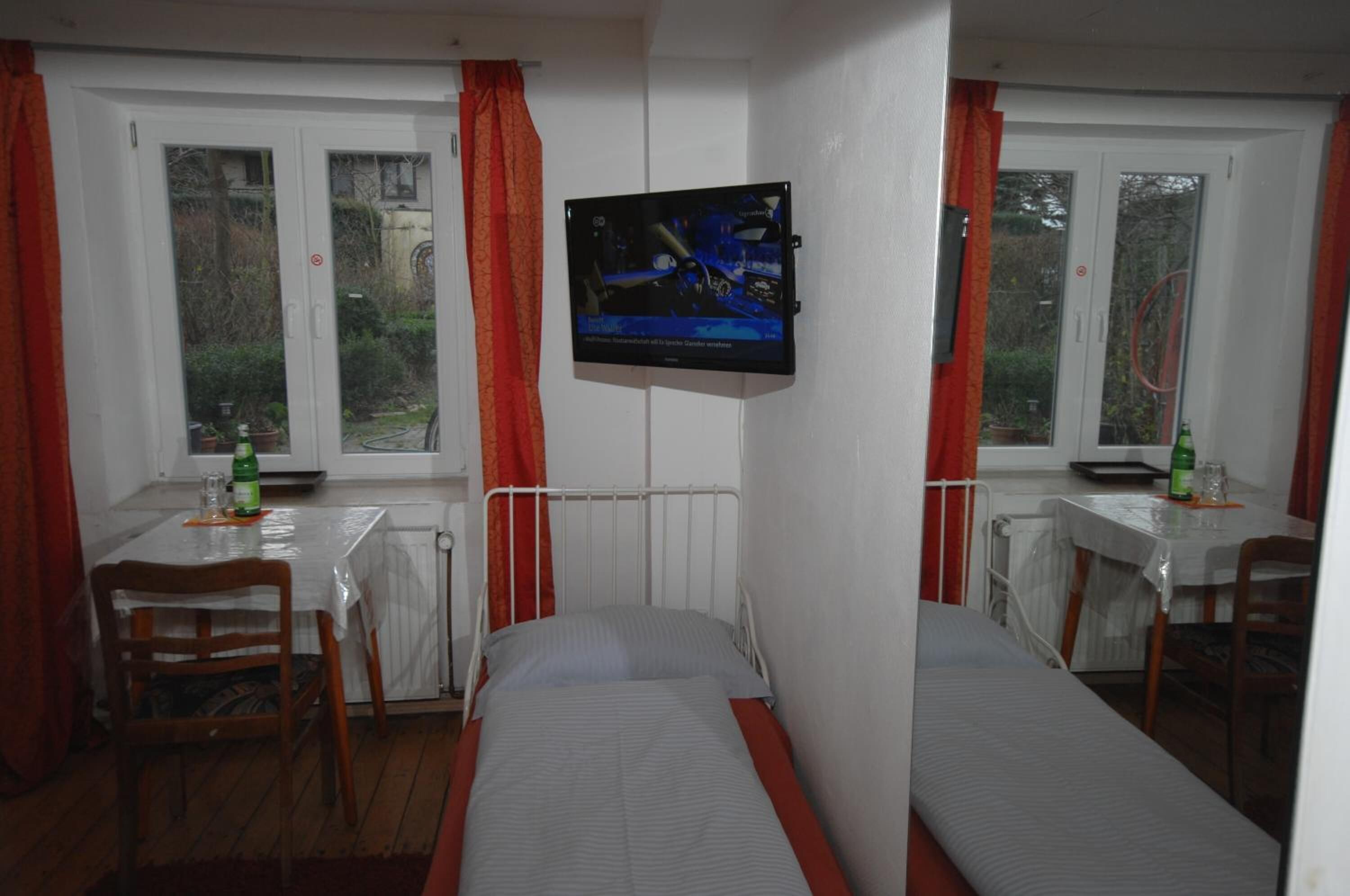 Ein kleines, sauberes Zimmer mit zwei Einzelbetten, einem Tisch mit Stühlen, einem an der Wand montierten Fernseher und Fenstern mit Vorhängen.